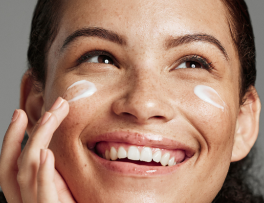 Smiling woman with freckles applying cream to her cheek.