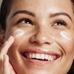 Smiling woman with freckles applying cream to her cheek.