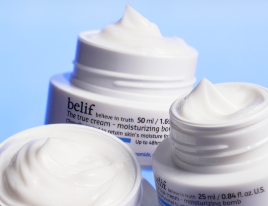 Three open jars of Belif "The true cream - moisturizing bomb" with peaks of white cream, on a light blue background.