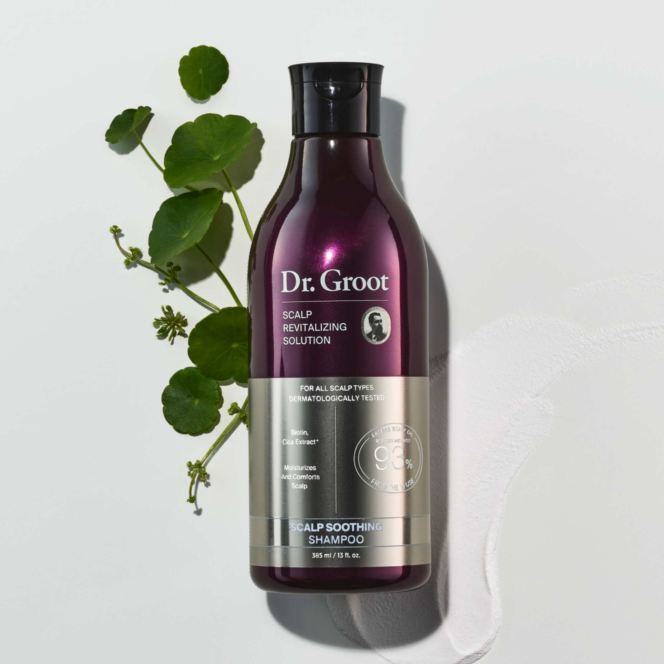 A dark purple bottle of Dr. Groot Scalp Soothing Shampoo lying on a grey surface in a white spread of the shampoo with a cica sprig.