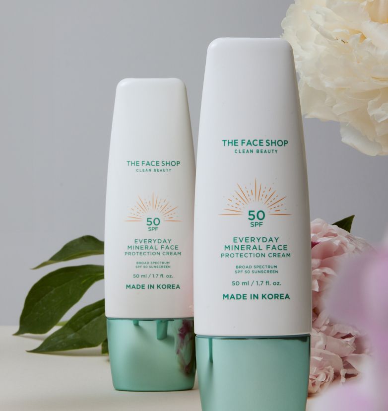 Two bottles of A bottle of The Face Shop Clean Beauty Everyday Mineral Face Protection Cream SPF 50. A leaf and two flowers are placed behind them.