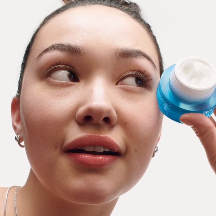 A model looking out of frame to the left, holding an open mini jar of belif Moisturizing Eye Bomb on a white background.