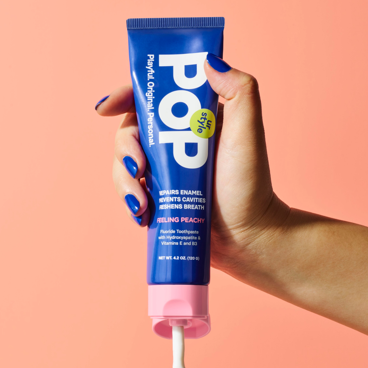A hand with blue-painted nails holding a blue tube of POP Feeling Peachy Toothpaste with a pink cap on a peach surface