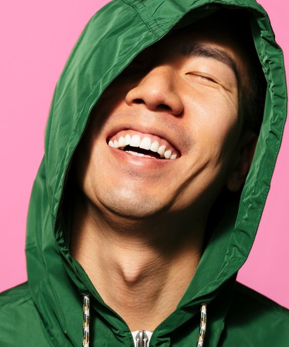 A young man wearing a green hoodie with the hood on, eyes closed and smiling on a light pink background.