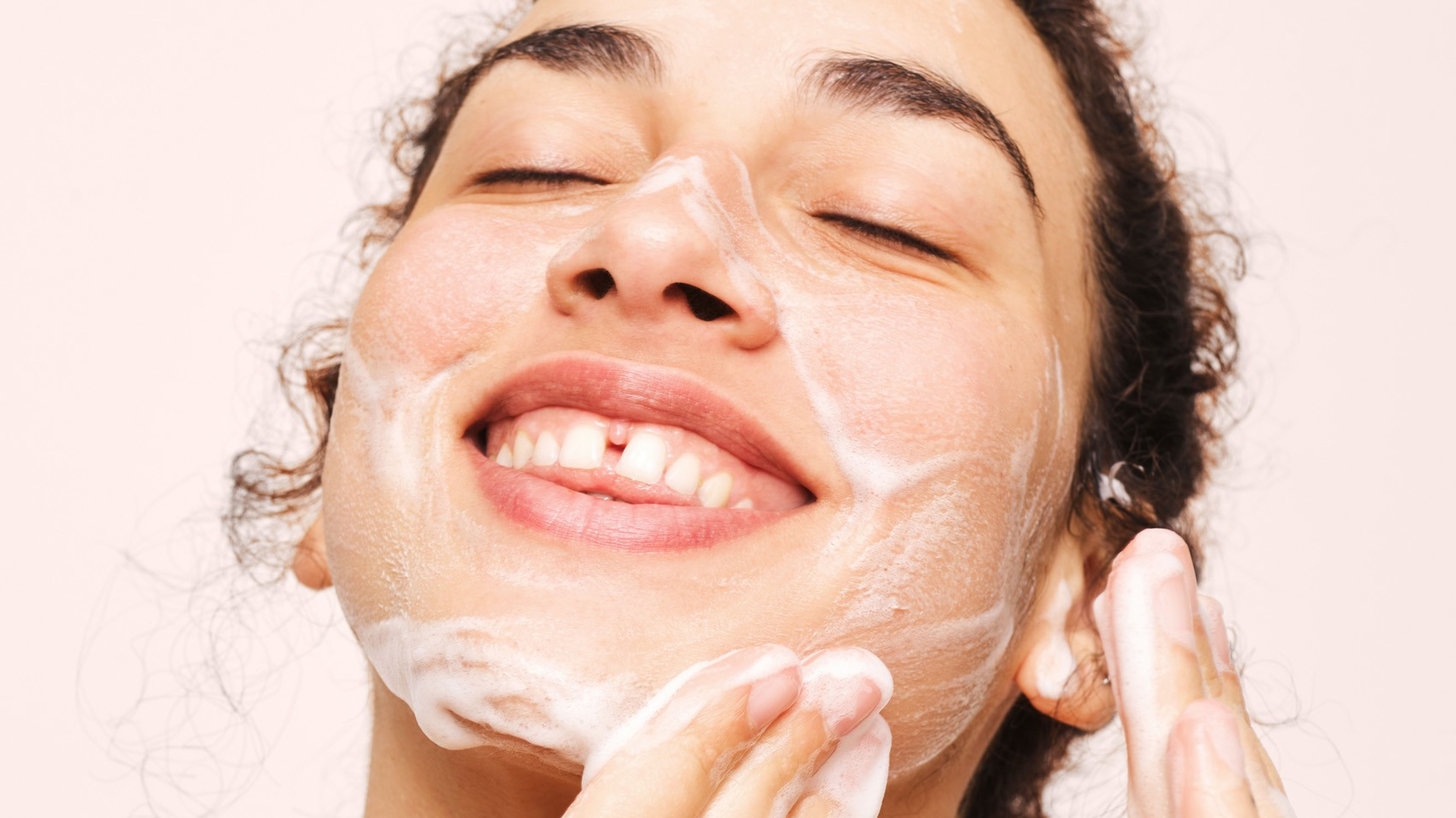 A smiling person with closed eyes happily washes their face with a foaming cleanser.