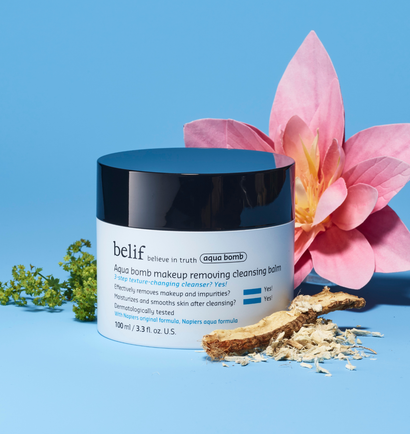 A white jar of belif Aqua Bomb Makeup Removing Cleansing Balm standing on a light blue surface among flowers and twigs