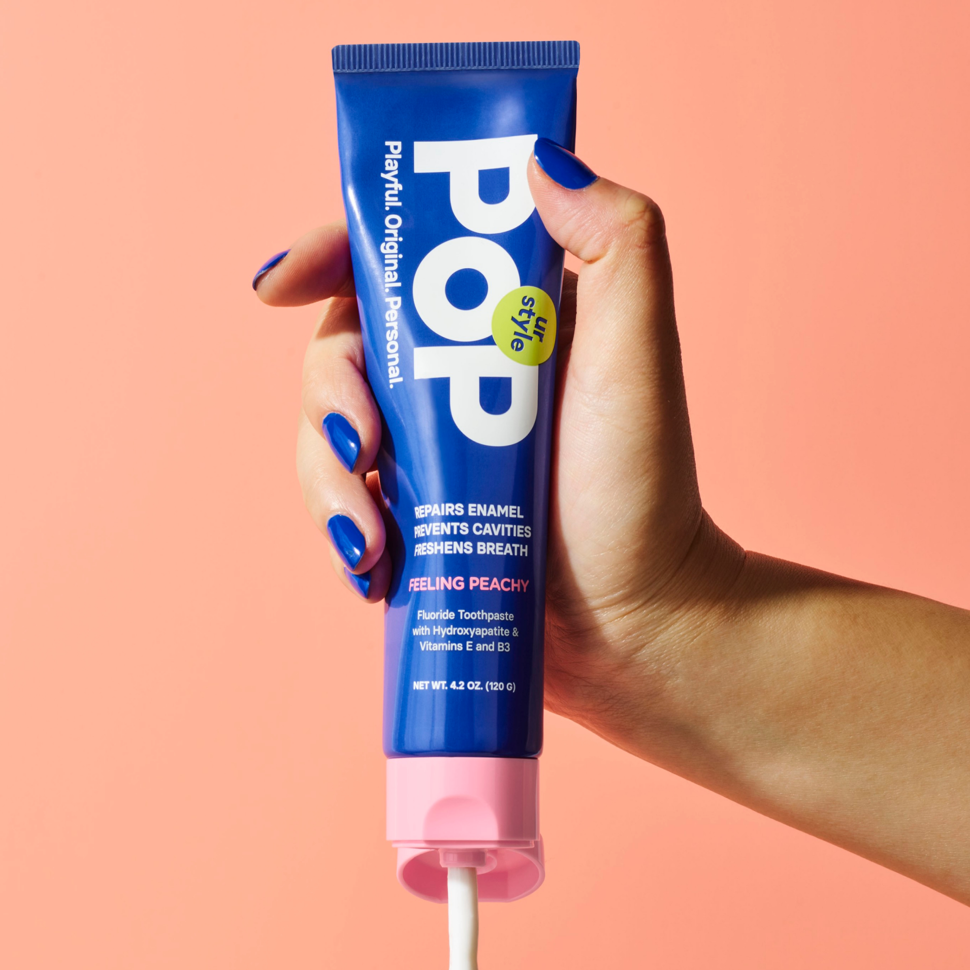 A hand with blue-painted nails holding a blue tube of POP Feeling Peachy Toothpaste with a pink cap on a peach surface