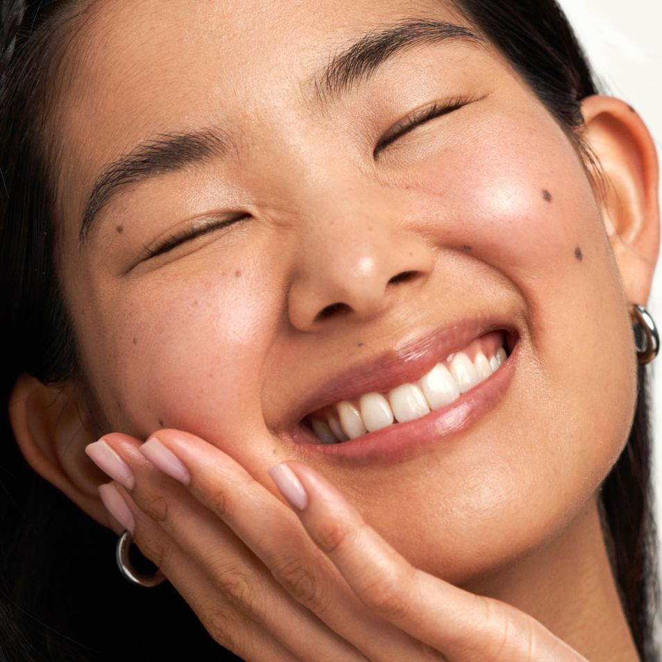 A woman with closed eyes smiles widely, her hand gently touching her cheek.