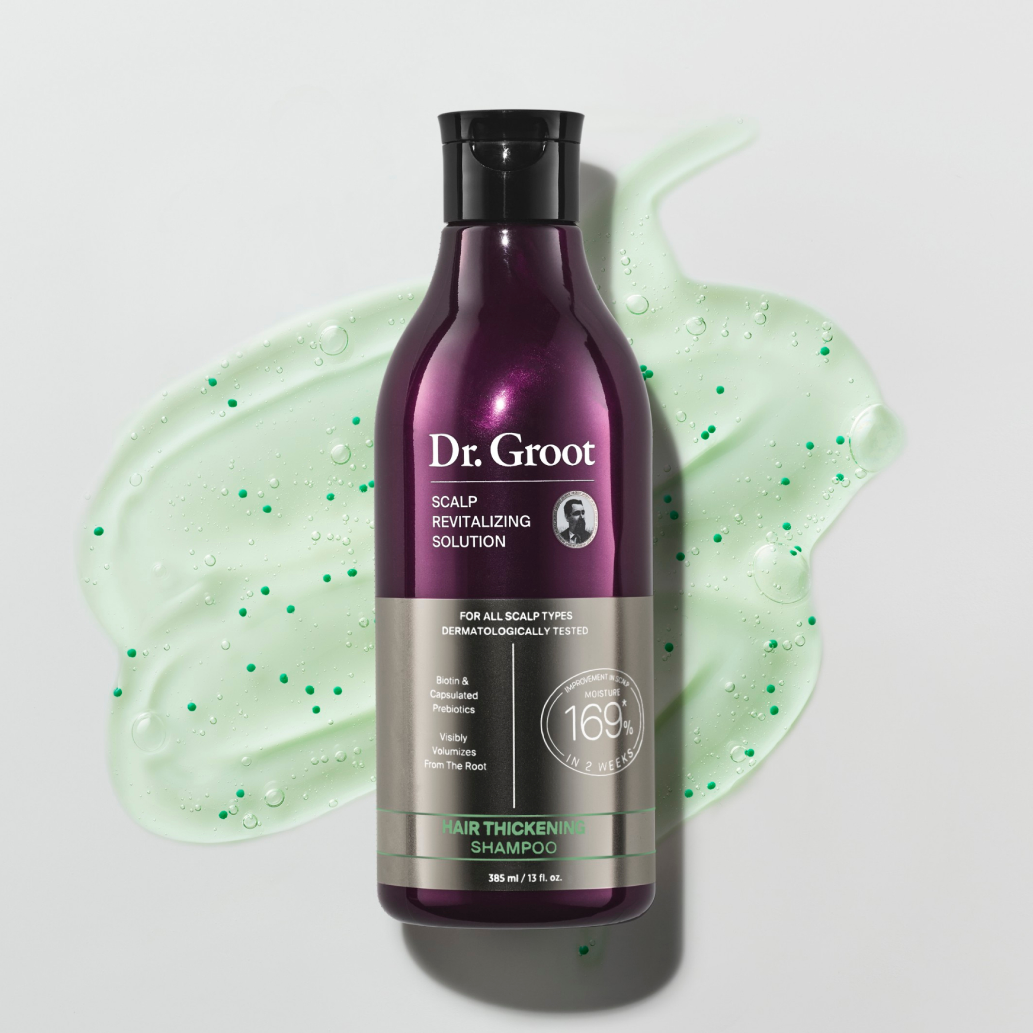 A dark purple bottle of Dr. Groot Hair Thickening Shampoo lying on a grey surface in a light green spread of the shampoo.