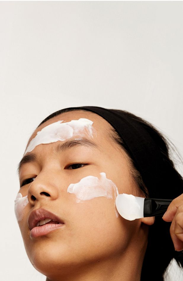 A closeup of a model applying skincare cream product to her face with a brush object.
