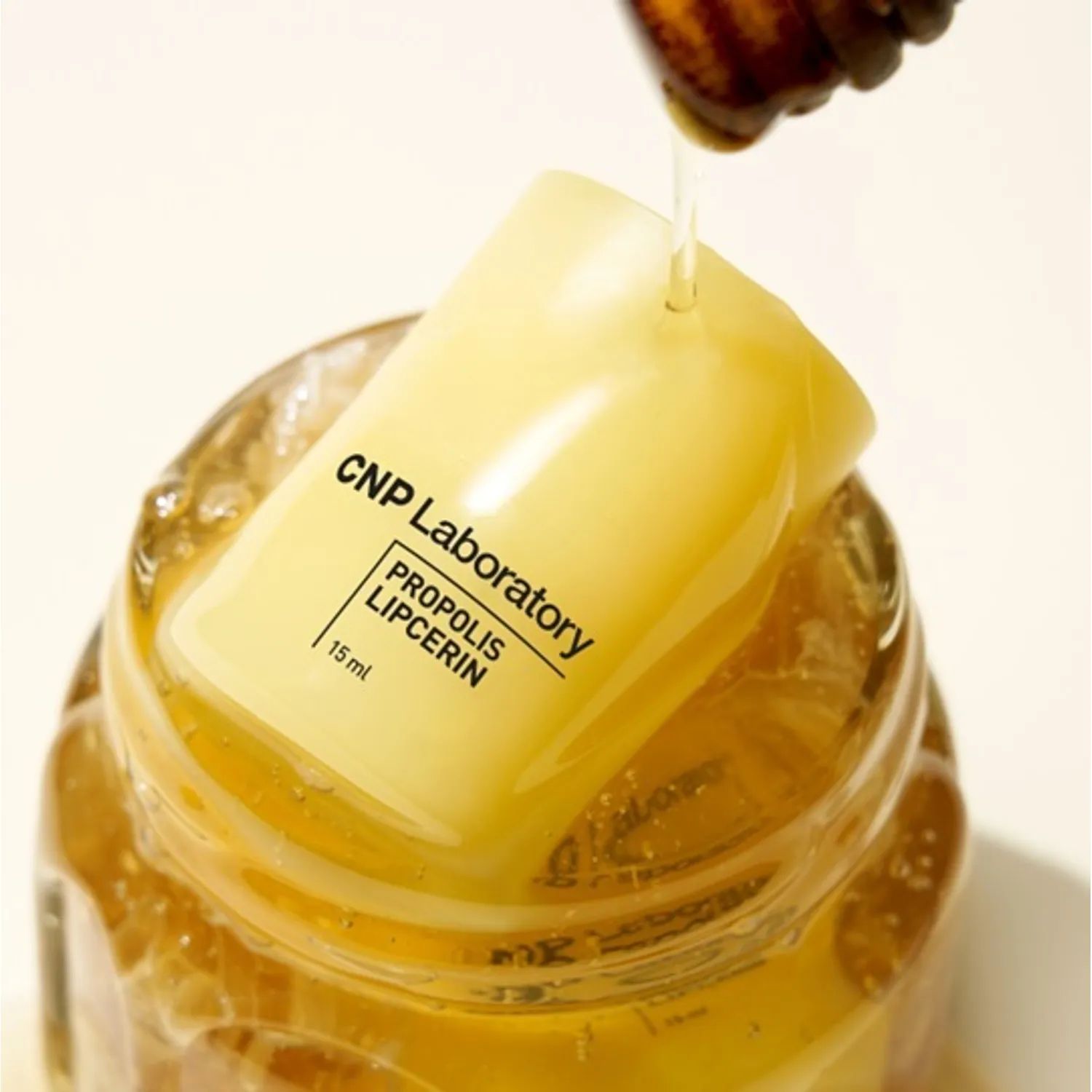 A yellow tube of CNP Laboratory Propolis Lipcerin partially submerged in honey, with a honey dipper pouring more honey onto it.
