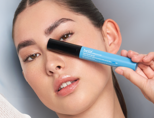 A woman holds a light blue tube of Belif Aqua Bomb eye gel across her face, covering the corner of one eye.