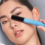 A woman holds a light blue tube of Belif Aqua Bomb eye gel across her face, covering the corner of one eye.
