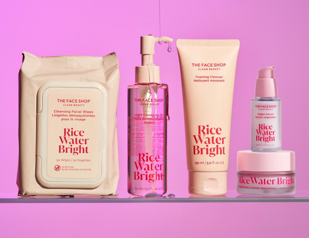 Five THE FACE SHOP Rice Water Bright skincare products: wipes, cleansing oil, foaming cleanser, serum, and cream, on a clear shelf against a pink background.