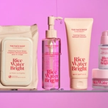 Five THE FACE SHOP Rice Water Bright skincare products: wipes, cleansing oil, foaming cleanser, serum, and cream, on a clear shelf against a pink background.