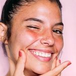 A model winking at the viewer, smiling with her teeth, holding her chin with her fingers.