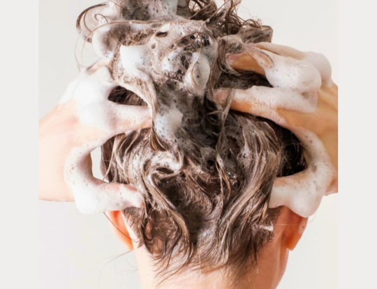 The back of a woman's hair with white shampoo lather in it and the woman's hands massaging the shampoo into the hair.