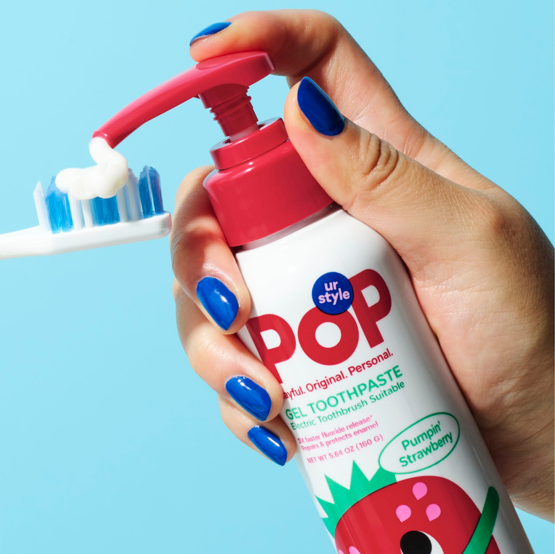 A hand holding a white bottle of POP Pumpin' Strawberry Gel Toothpaste, pumping the toothpaste out on a white-and-blue toothbrush ona a light blue background.