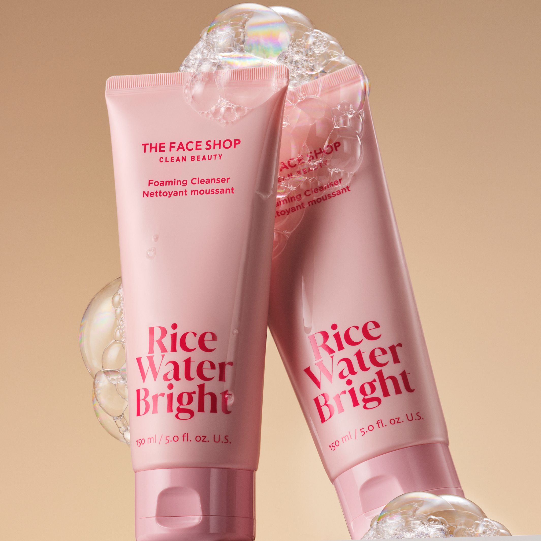 Two tubes of the face shop rice water bright cleansing foam covered in bubbles is displayed on a warm beige background.