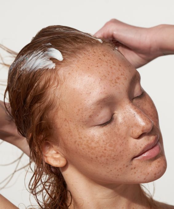 Close-up of a person with freckles and closed eyes, having a white product massaged into their wet, reddish hair.