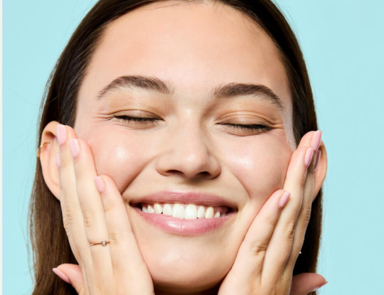 Smiling woman with closed eyes touching her radiant face against a light green background.