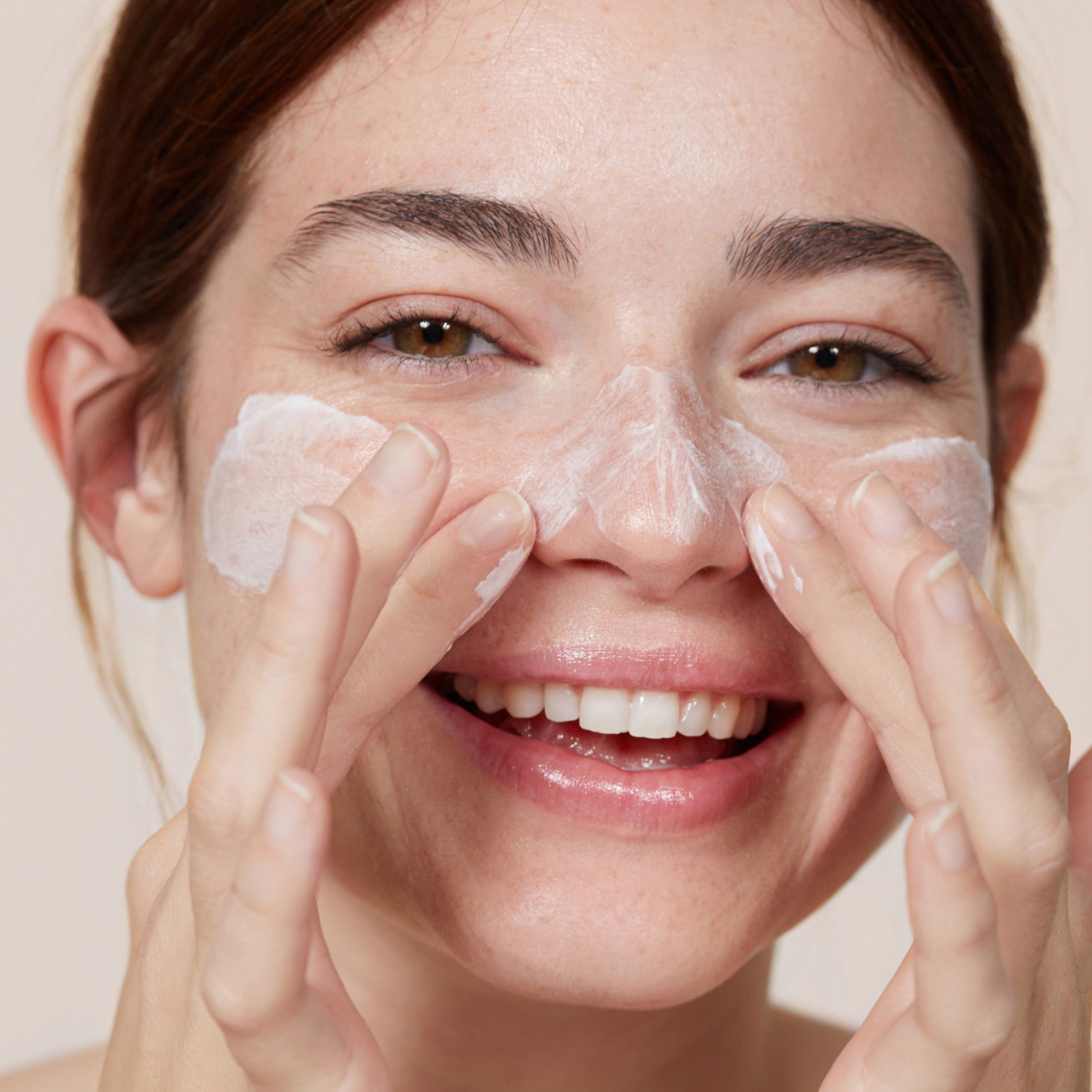Close up of a model smiling and rubbing cleanser on her face.