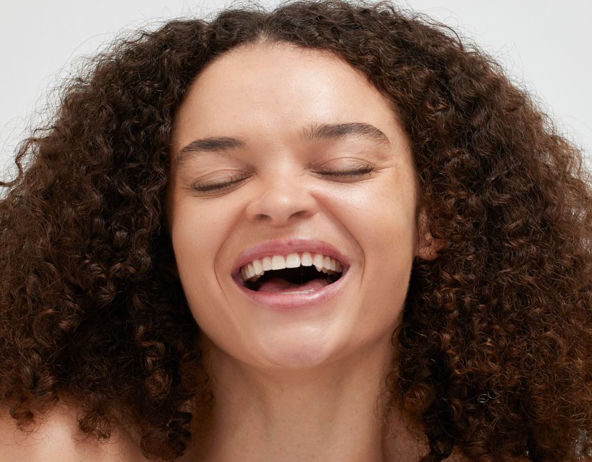 A person with curly hair is laughing joyfully with closed eyes and an open mouth. The background is plain and light-colored, emphasizing the person's happy expression.