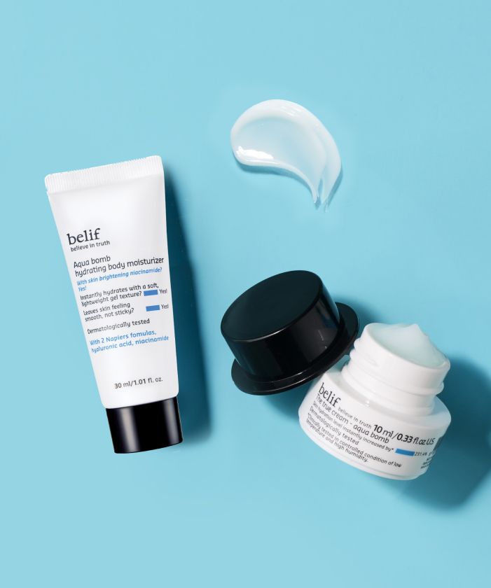Two skin care products by belif on a light blue background: an open jar of Aqua Bomb True Cream showing its white contents and a tube of Aqua Bomb body moisturizer with a dab of cream beside it.