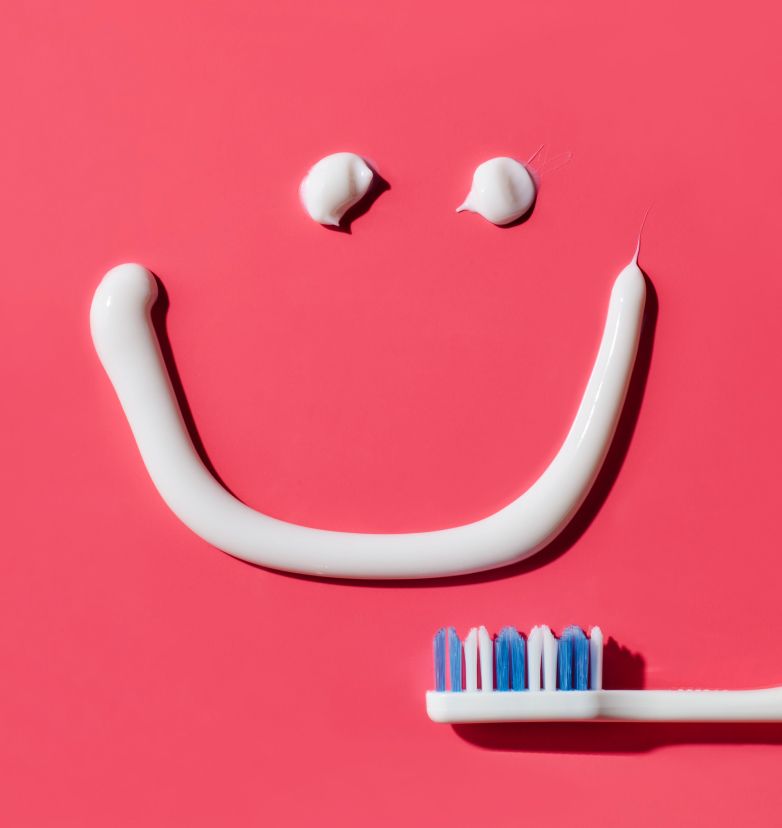 A white-and-blue toothbrush lying on a pink surface with a smiley face drawn with POP Kids Gel Pumpin' Strawberry Toothpaste above it