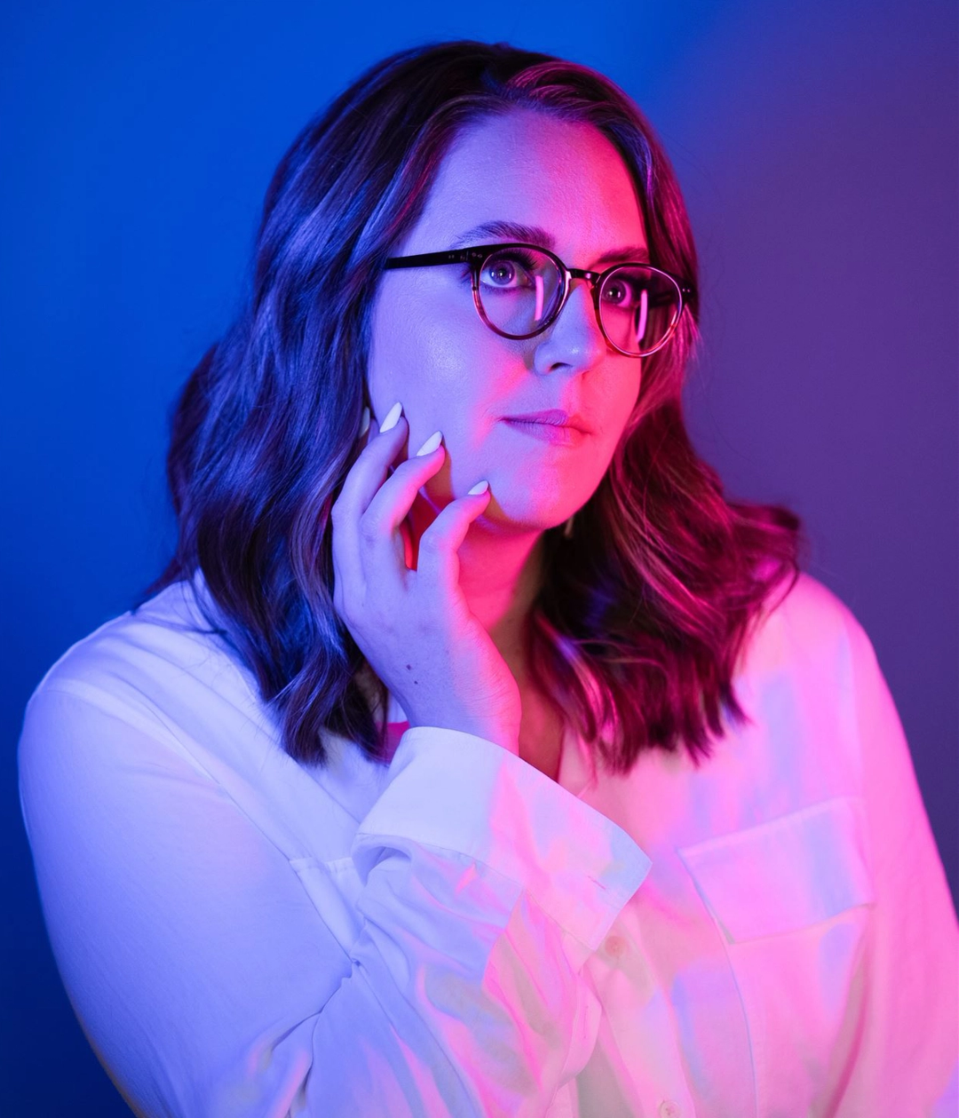 Professional headshot of Alecia, featuring a vibrant pink and blue lighting effect. Alecia is wearing glasses and a white shirt, with shoulder-length hair and a confident, contemplative expression, gently resting a hand near their face.