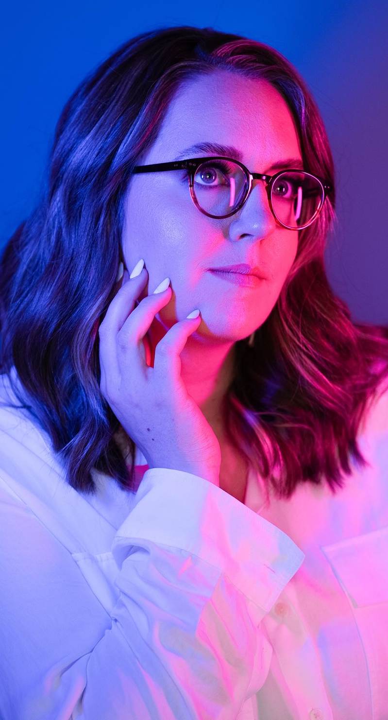 Professional headshot of Alecia, featuring a vibrant pink and blue lighting effect. Alecia is wearing glasses and a white shirt, with shoulder-length hair and a confident, contemplative expression, gently resting a hand near their face.