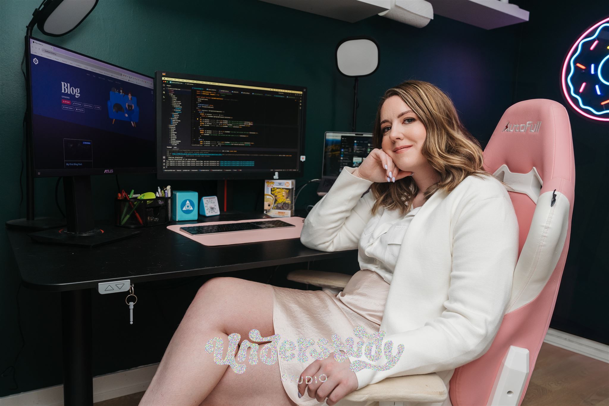 Alecia, a smiling woman, sits at a desk with a dual-monitor setup, actively coding on one screen while a blog page is displayed on the other. She is seated in a pink gaming-style chair and looks back toward the camera. The workspace is neatly arranged with small desk items.