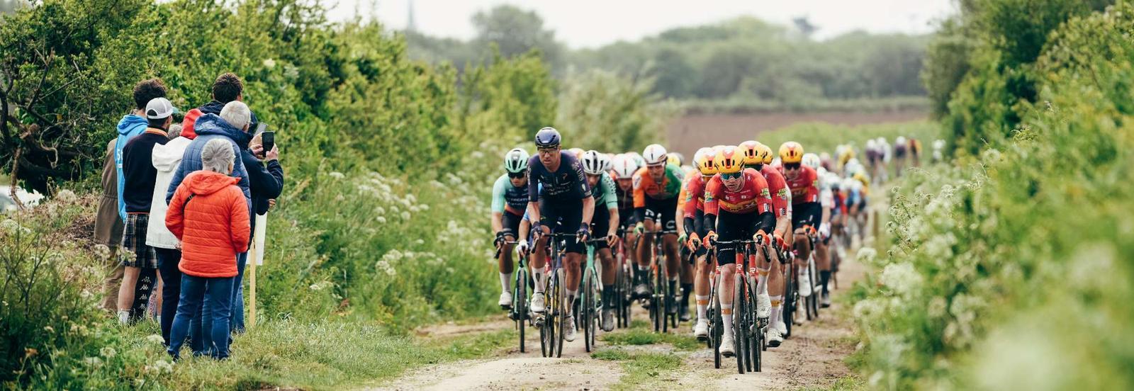 Riders crossing a hedge-lined Breton gravel farm track (ribin) with spectators watching from the roadside