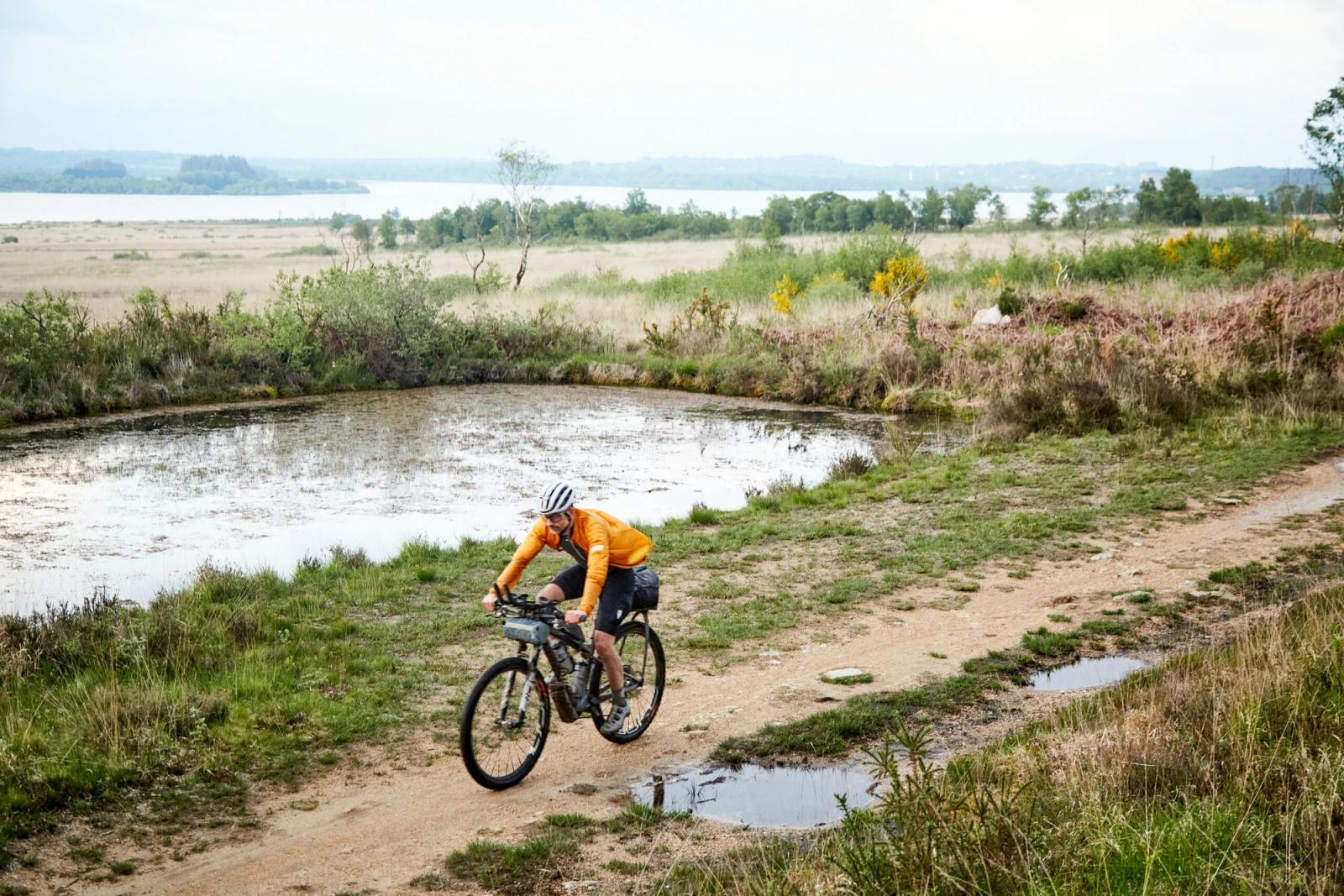 Gravel rider crossing a wetland track beside a lake in rural Brittany