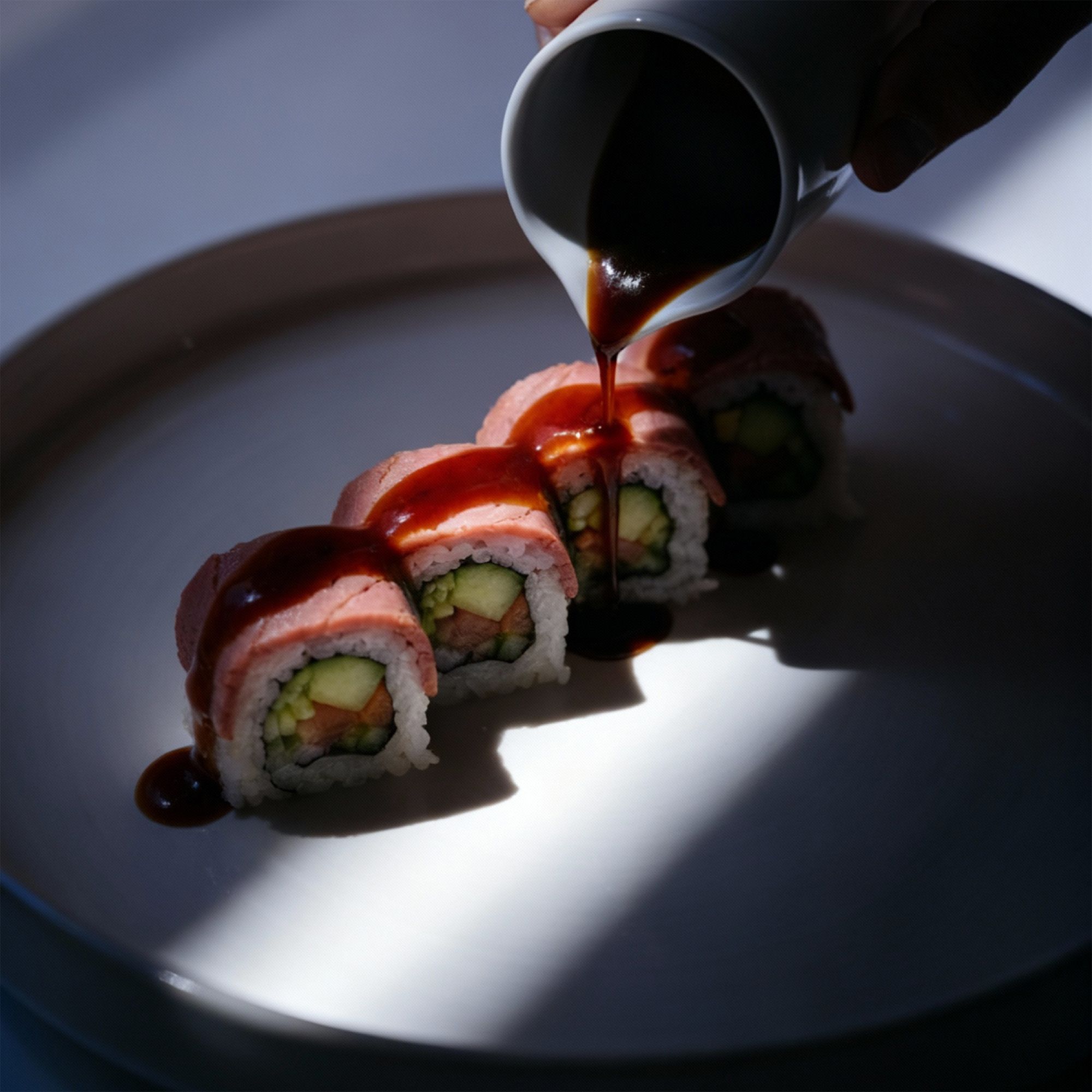 Sushi on a white plate with a hand pouring sauce on top on a white table