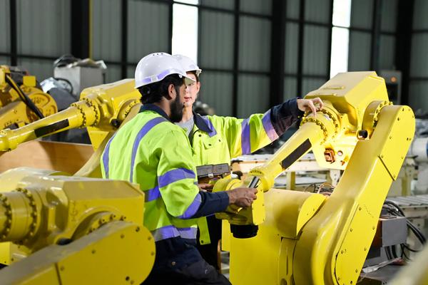 Two industrial workers in hi-vis gear operating yellow robotic arms on a production line using a teach pendant