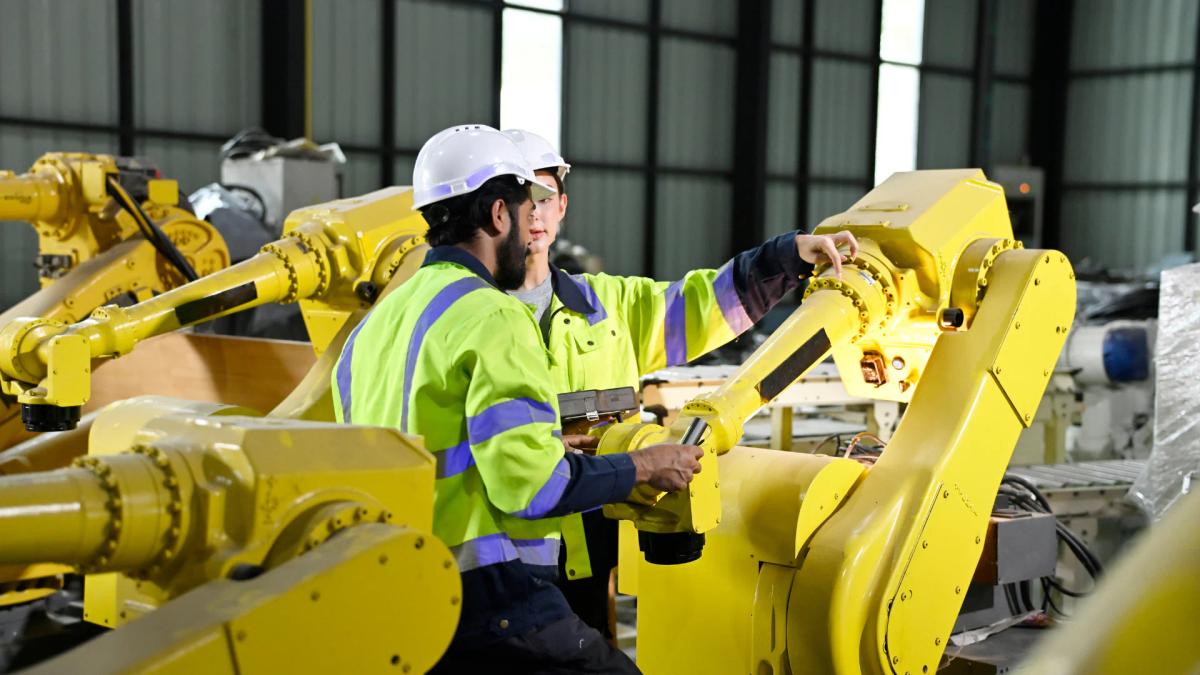 Two industrial workers in hi-vis gear operating yellow robotic arms on a production line using a teach pendant