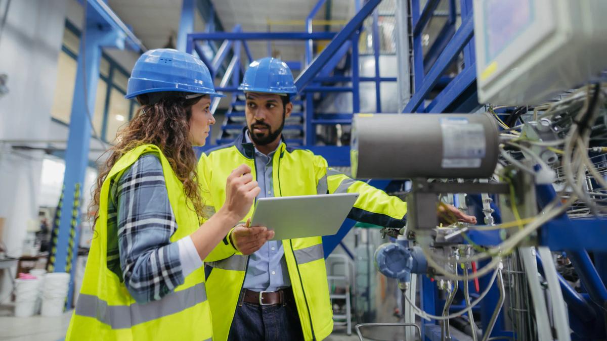 Two engineers in hard hats and hi-vis vests reviewing equipment data on a tablet on the plant floor