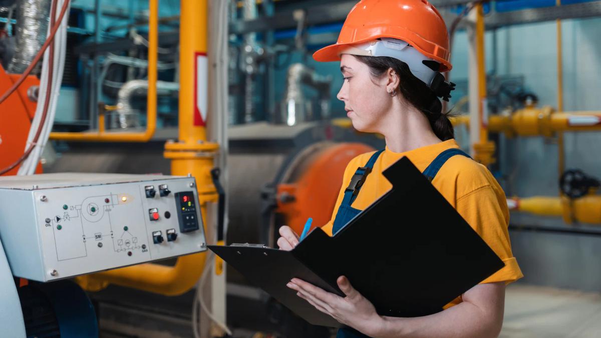 Engineer in an orange hard hat recording equipment readings on a clipboard next to an industrial control panel