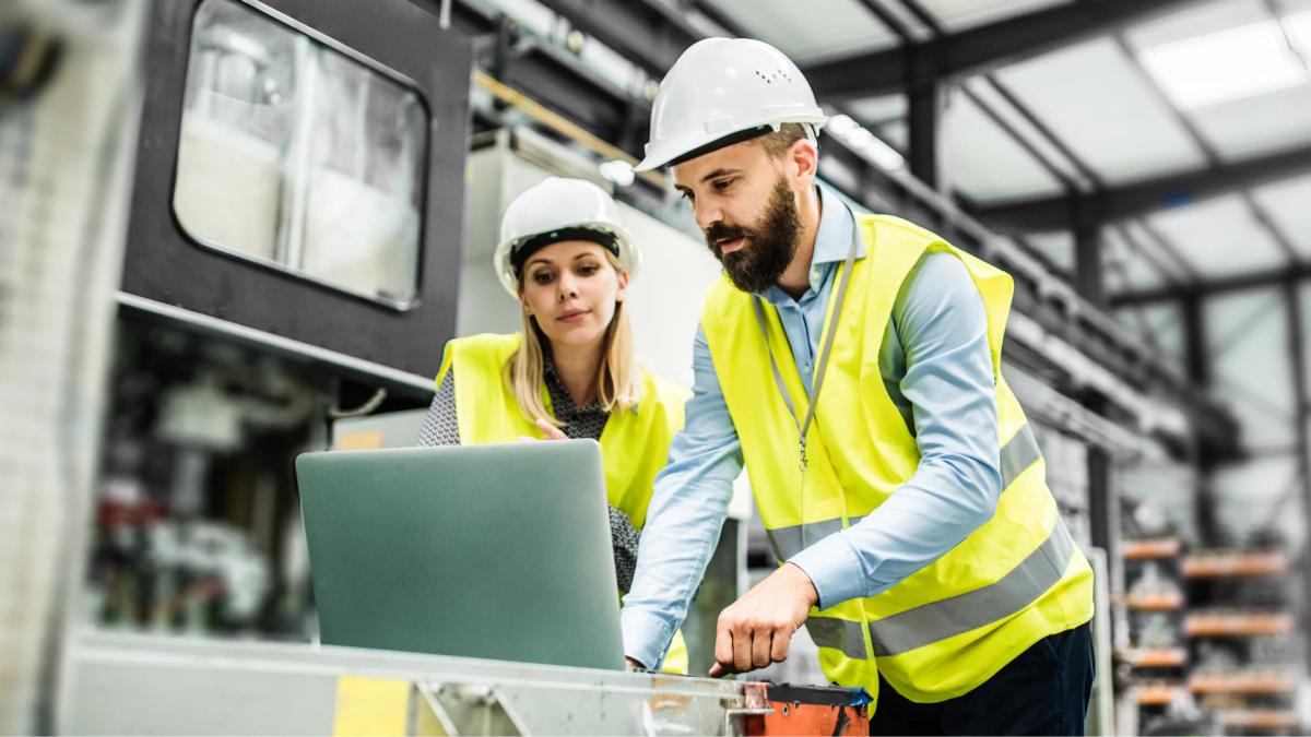 Two engineers in hard hats and hi-vis vests reviewing data on a laptop next to industrial equipment