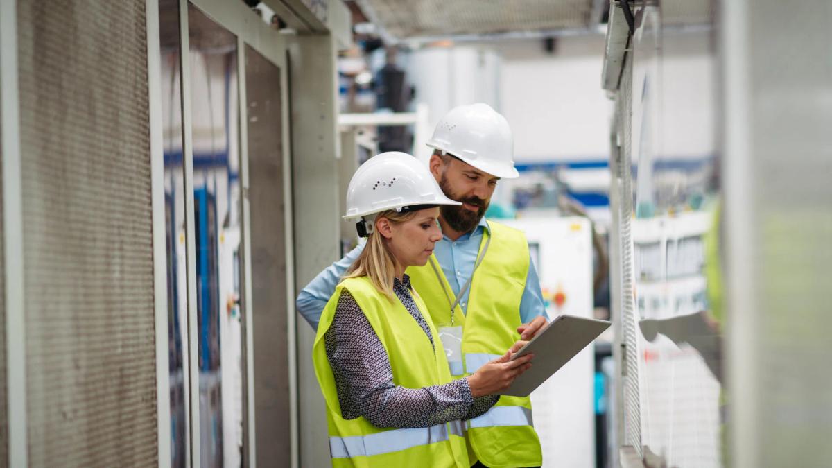 Two engineers in hard hats and hi-vis vests reviewing an electrical panel using a tablet