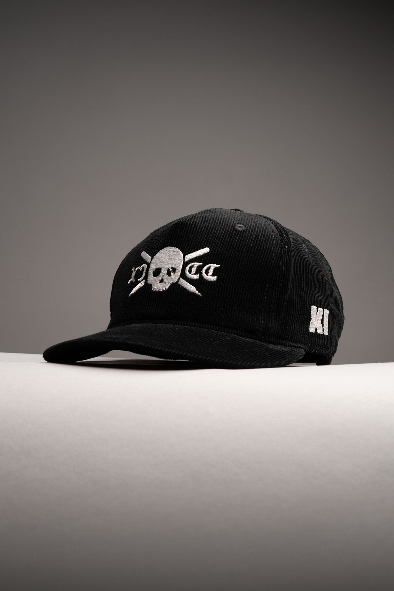 Black Eleven Cricket Club cap with a white skull-and-crossed-bats emblem on a grey studio surface.