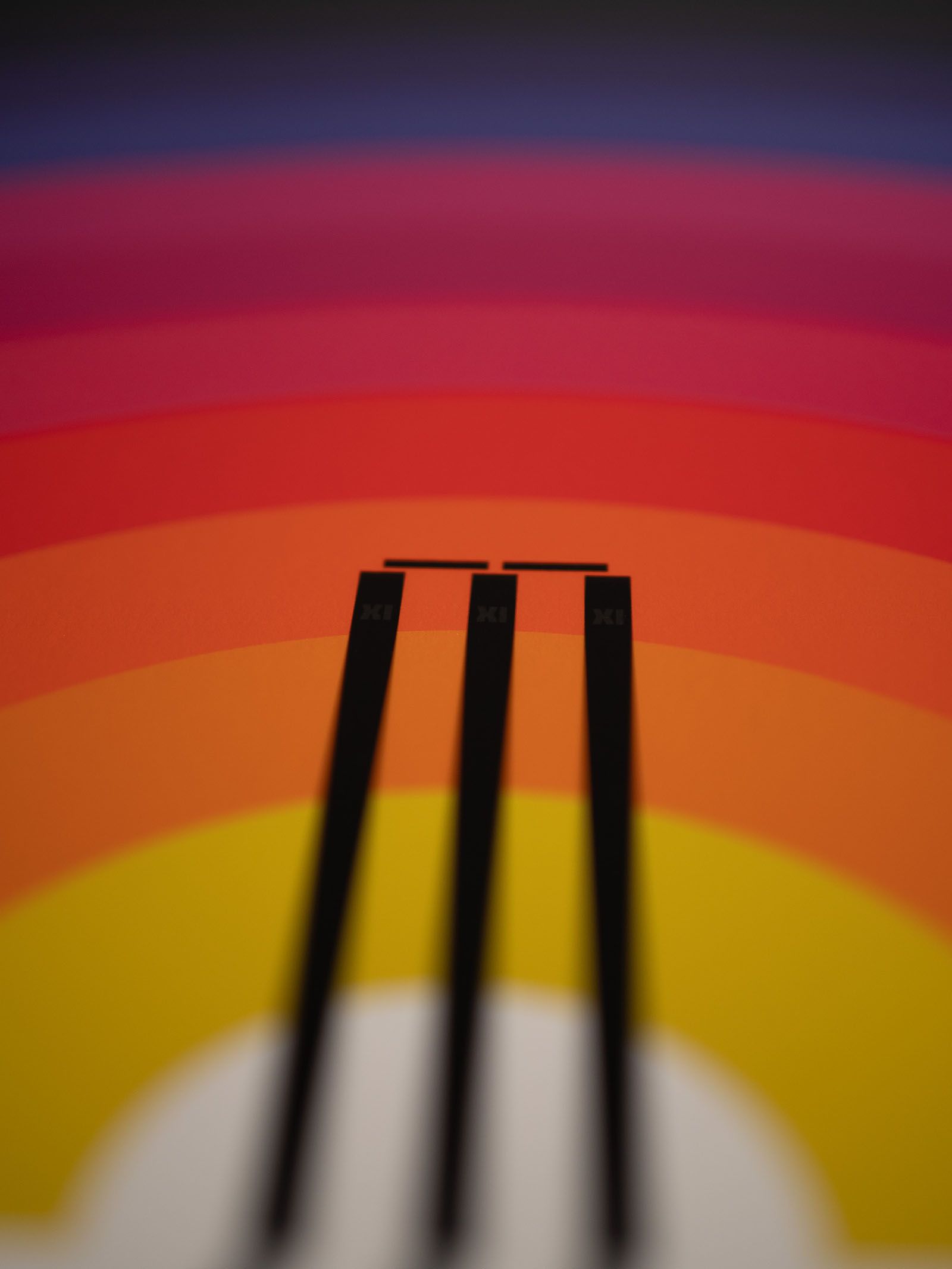 A cricket-stump silhouette rising through bands of yellow, orange, red and purple.