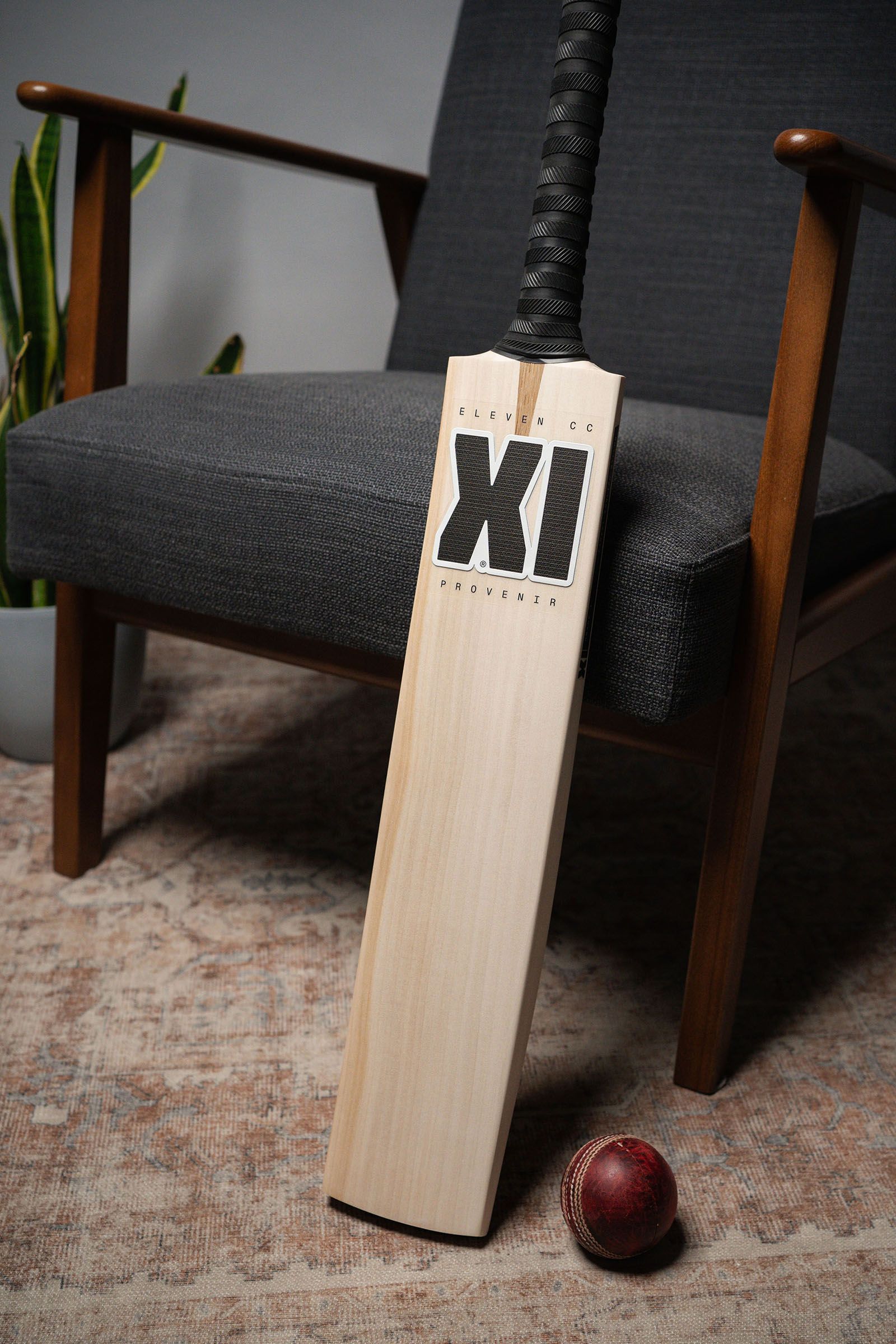 An XI-branded cricket bat leaning against a wooden armchair beside a red cricket ball.