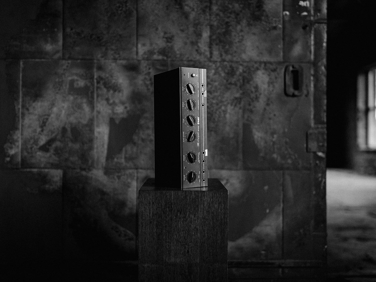 A tall audio equipment enclosure on a pedestal against a textured studio wall, photographed in black and white.