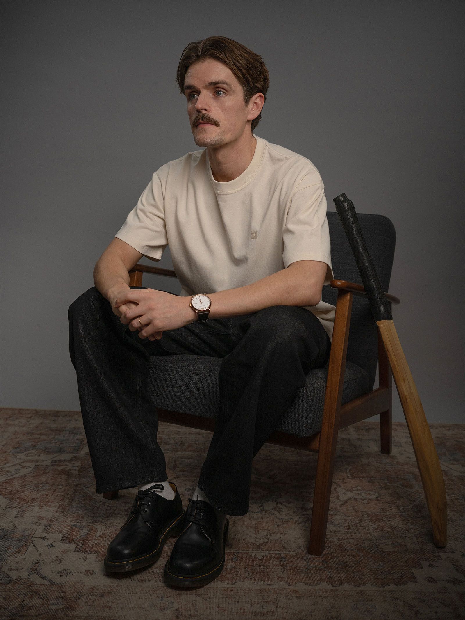 A model in a cream Eleven Cricket Club T-shirt seated in a wooden armchair.