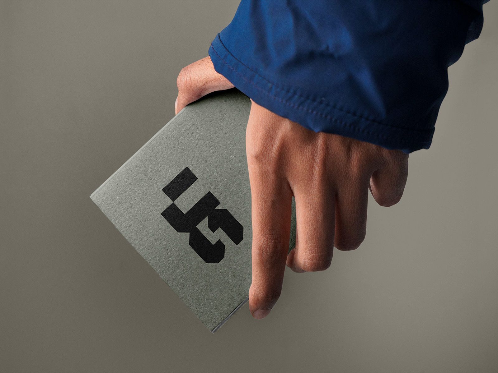 A hand holding a grey notebook with a bold black geometric monogram.