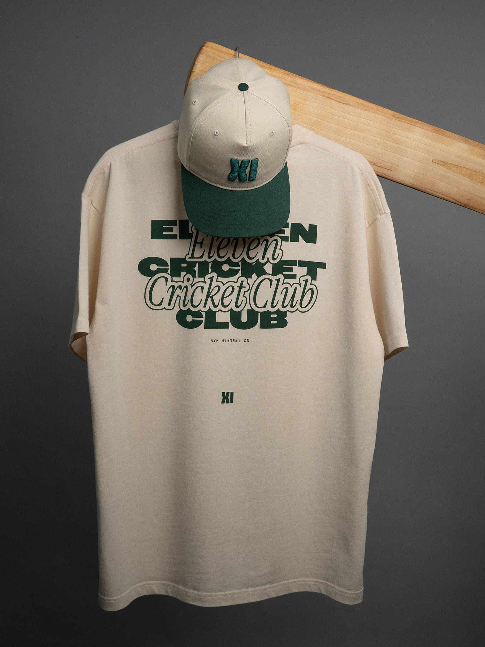 Cream Eleven Cricket Club T-shirt and cream-and-green cap displayed on a wooden hanger.