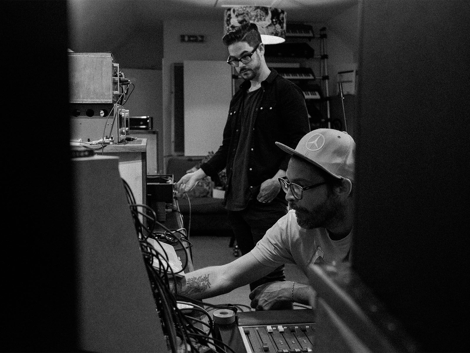 Two people working with electronic instruments in a recording studio, photographed in black and white.