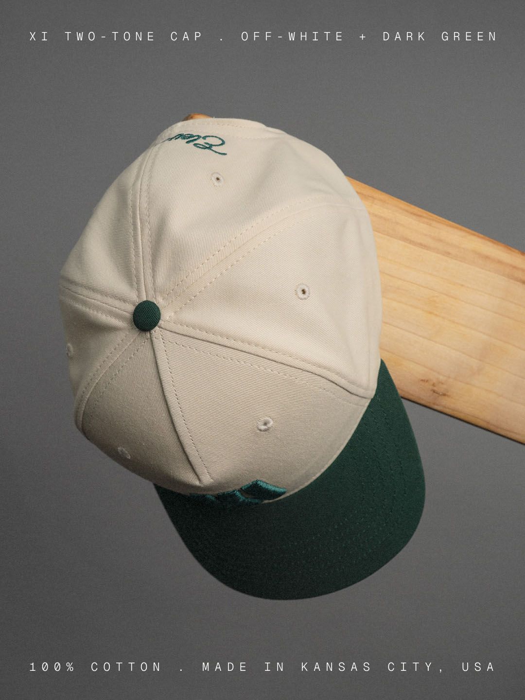 Cream-and-green Eleven Cricket Club cap hanging from a wooden bat against a grey background.